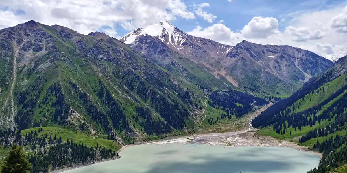 Big Almaty Lake in summer, with spruce-covered green slopes and a snow-capped peak of the Ile Alatau behind it
