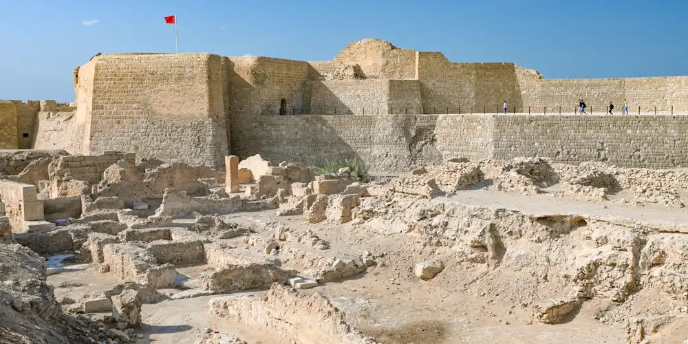 Stone walls of Qal'at al-Bahrain above excavated ancient ruins, with a Bahraini flag, under a clear sky