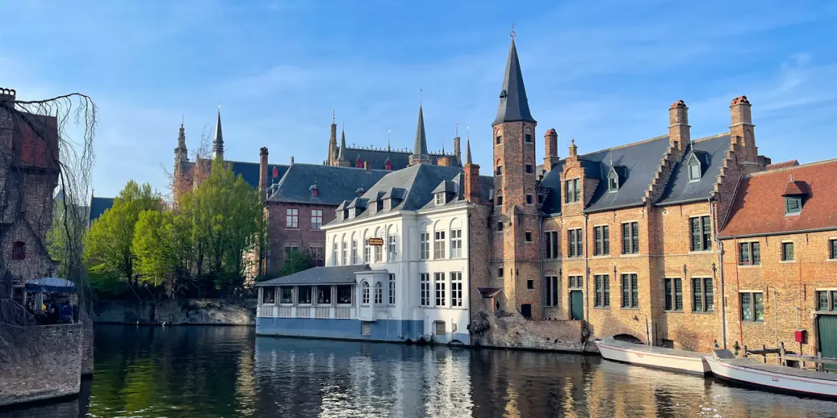 Brick and white-walled houses and a slender spired tower rising straight out of a still canal in Bruges, with canal boats moored on the right
