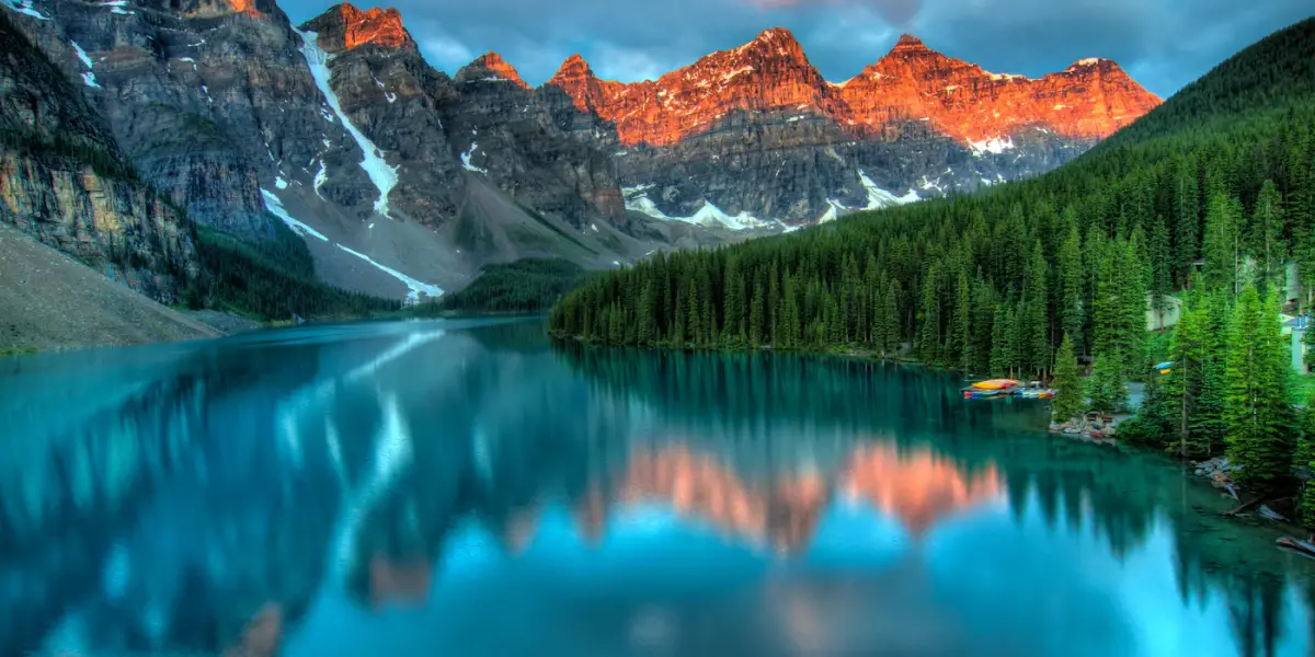 Moraine Lake in Banff National Park at sunrise, the orange-lit peaks of the Valley of the Ten Peaks reflected in the turquoise water, with canoes moored below the forest on the right