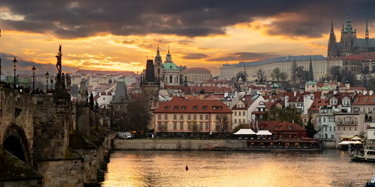 Charles Bridge at sunset seen from the Old Town bank of the Vltava, with the green dome of St Nicholas in the Lesser Town and St Vitus Cathedral at Prague Castle on the hill behind
