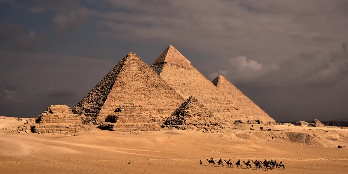 A line of camels crossing the sand in front of the three pyramids of Giza and the small queens' pyramids beside them, under a stormy sky