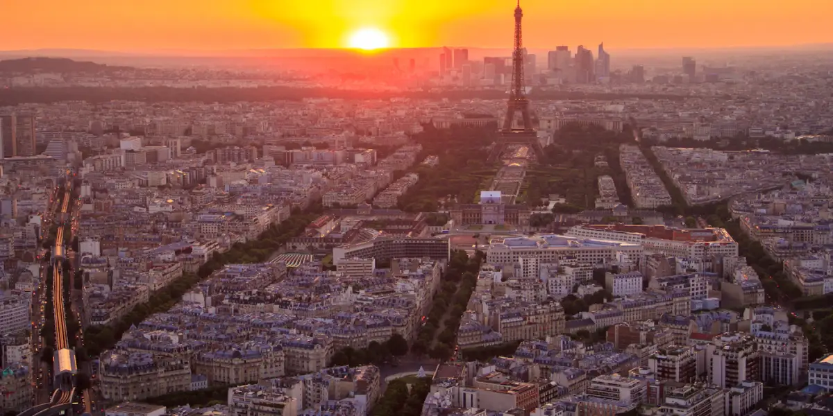The Eiffel Tower and the Champ de Mars at sunset, seen from high above the rooftops of Paris, with a cluster of high-rise towers on the horizon