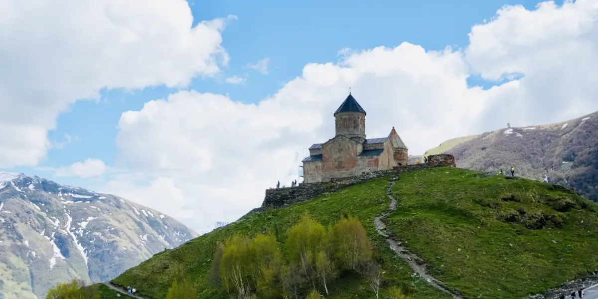 Gergeti Trinity Church on its green hilltop above Stepantsminda, with snow-streaked Caucasus slopes and clouds behind