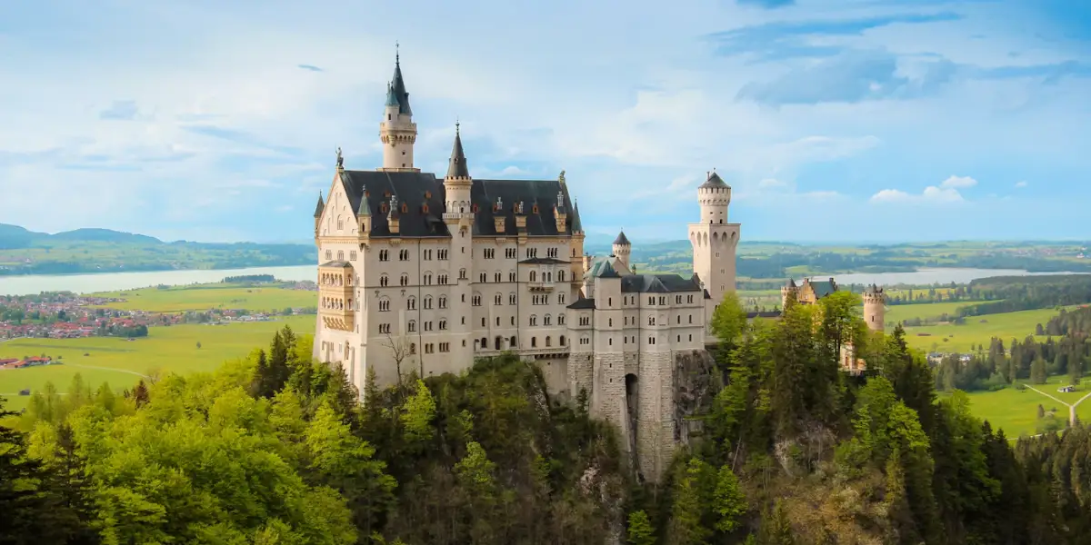 Neuschwanstein Castle on its forested crag, with green farmland and a lake stretching away behind it