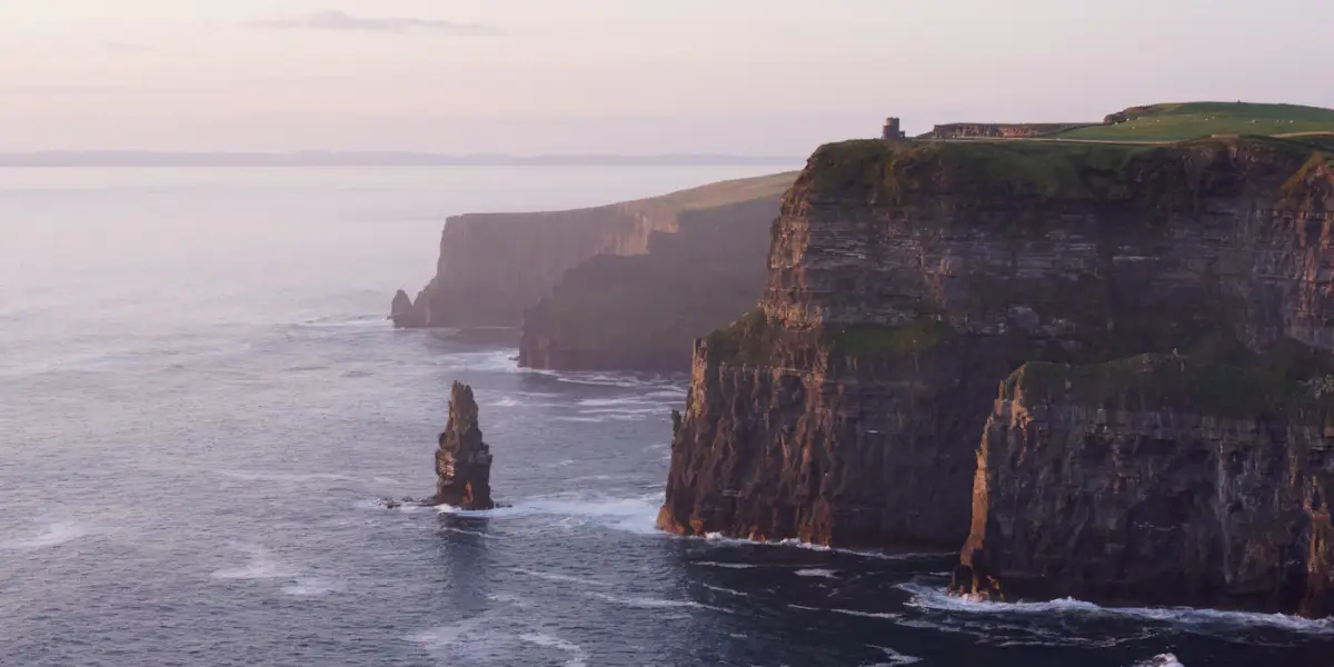 The Cliffs of Moher in soft pink light, with O'Brien's Tower on the cliff top at right, a sea stack in the Atlantic below and more cliffs fading into the haze