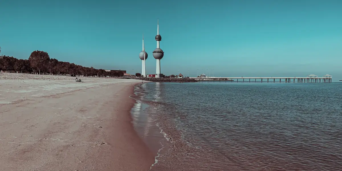 The Kuwait Towers rising beyond a sandy beach on the Kuwait City seafront, with a long pier running out over the calm Gulf water