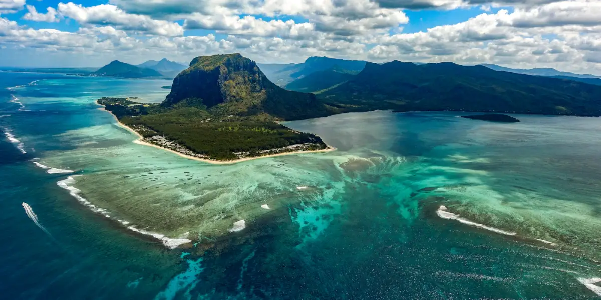 Aerial view of Le Morne Brabant on the south-west tip of Mauritius, its forested peninsula ringed by a turquoise lagoon, with the sand patterns of the 'underwater waterfall' spreading across the reef in the foreground