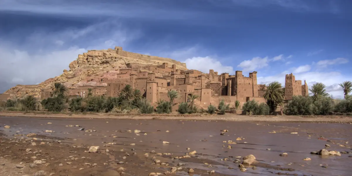 The mud-brick ksar of Aït-Benhaddou climbing its hill behind palm trees, seen across the shallow, stony bed of the Ounila River