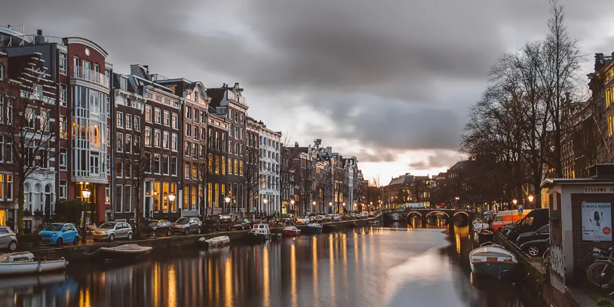An Amsterdam canal at dusk, with lit canal houses reflected in the water and an arched bridge in the distance