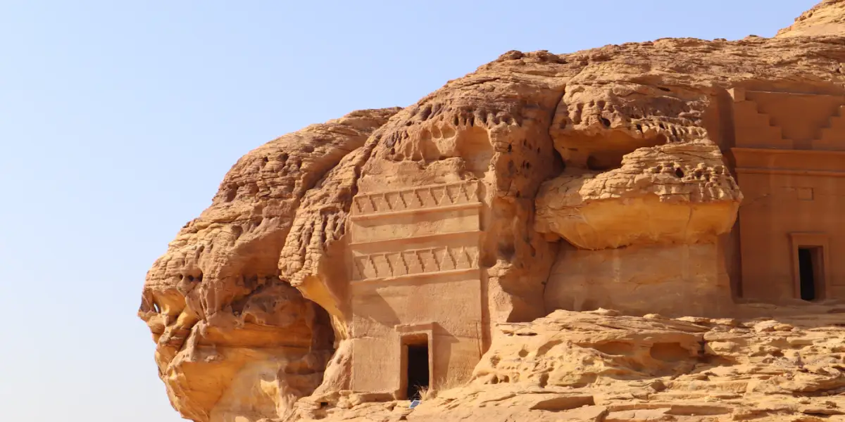 Nabataean tomb facades with carved stepped crowns and doorways cut into a weathered sandstone outcrop at Hegra, AlUla, under a clear sky