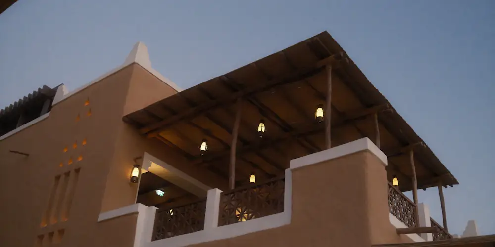 A mud-plastered Najdi-style building in Diriyah with a wooden-roofed balcony lit by lanterns at dusk