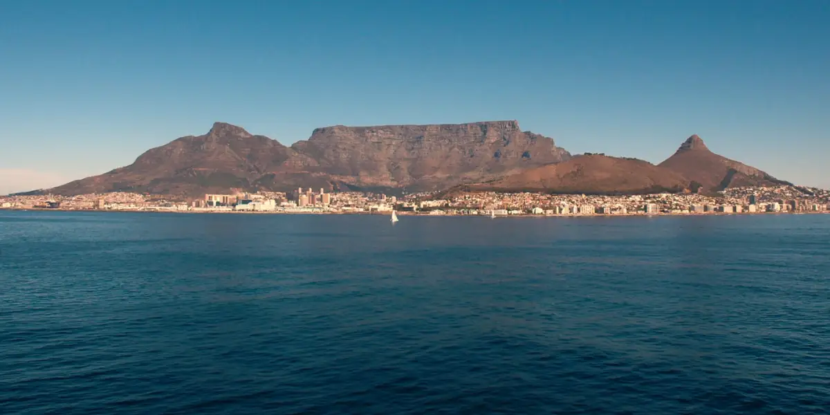 Table Mountain, with Devil's Peak on the left and Lion's Head on the right, rising behind the Cape Town waterfront, seen across the calm water of Table Bay with sailing boats