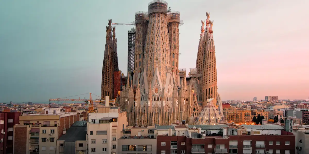 The towers of the Sagrada Família, with scaffolding and a crane on the central spires, rising above Barcelona's apartment blocks at dusk