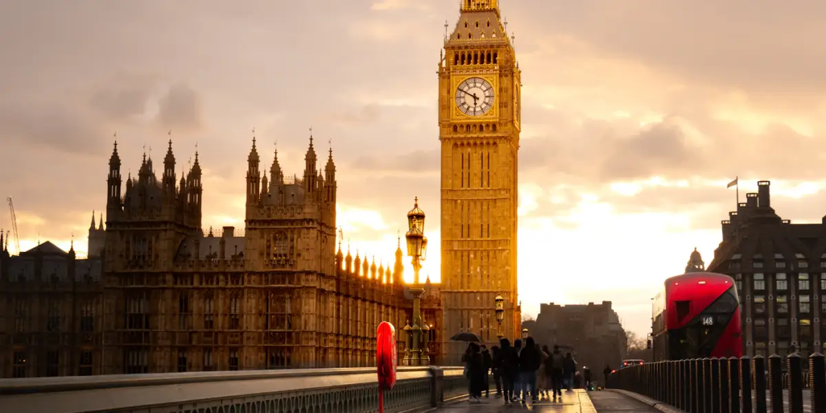 The Elizabeth Tower (Big Ben) and the Houses of Parliament lit gold at sunset, seen from Westminster Bridge with a red double-decker bus on the right